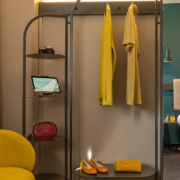 Une chambre d’hôtel moderne conçue par GH & Partners met en valeur L’Innovation durable chez AccorInvest. L’élégant présentoir en métal présente des vêtements jaunes, un appareil photo, une tablette, un sac rouge et des chaussures jaunes. À proximité se trouvent une chaise jaune et un lit avec une tête de lit beige. Les murs neutres et verts ajoutent de la chaleur à cet intérieur circulaire d’Ibis.