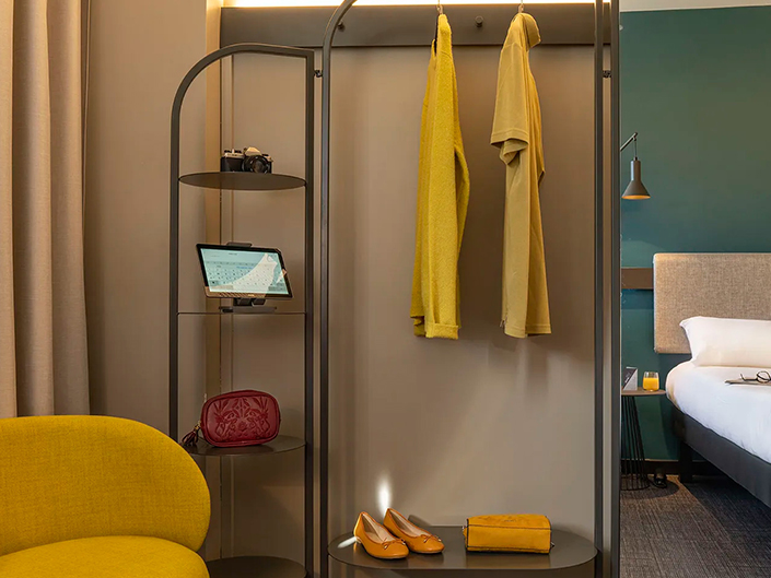 Une chambre d’hôtel moderne conçue par GH & Partners met en valeur L’Innovation durable chez AccorInvest. L’élégant présentoir en métal présente des vêtements jaunes, un appareil photo, une tablette, un sac rouge et des chaussures jaunes. À proximité se trouvent une chaise jaune et un lit avec une tête de lit beige. Les murs neutres et verts ajoutent de la chaleur à cet intérieur circulaire d’Ibis.