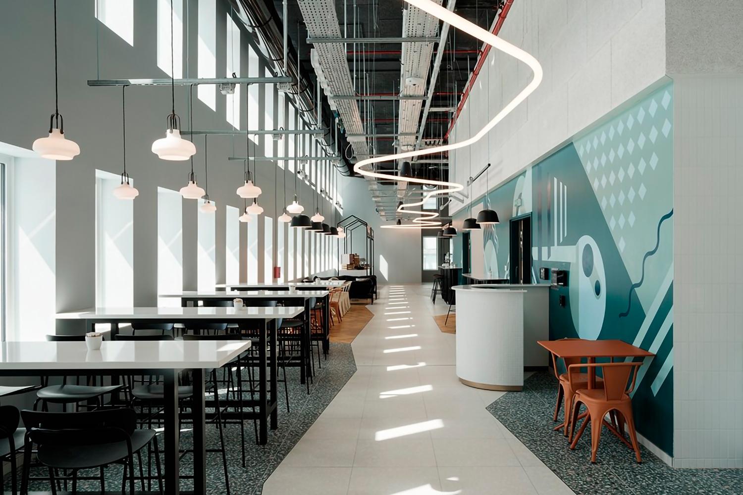 Un café moderne et spacieux avec de hauts plafonds et de grandes fenêtres laissant entrer une abondante lumière naturelle. Ibis_KAI présente un mélange de tables blanches et noires, des luminaires suspendus et un long luminaire incurvé. Les murs sont ornés de peintures murales géométriques dans des tons de bleu et de blanc.