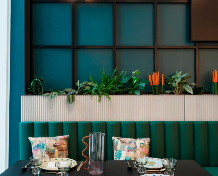 Une salle à manger moderne comprend une table dressée pour deux personnes avec des assiettes décoratives et des couverts. Derrière la table se trouve un banc vert rembourré orné de coussins fleuris. Une composition végétale et une suspension d'Ibis_KAI rehaussent l'ambiance sur un mur lambrissé bleu sarcelle foncé.