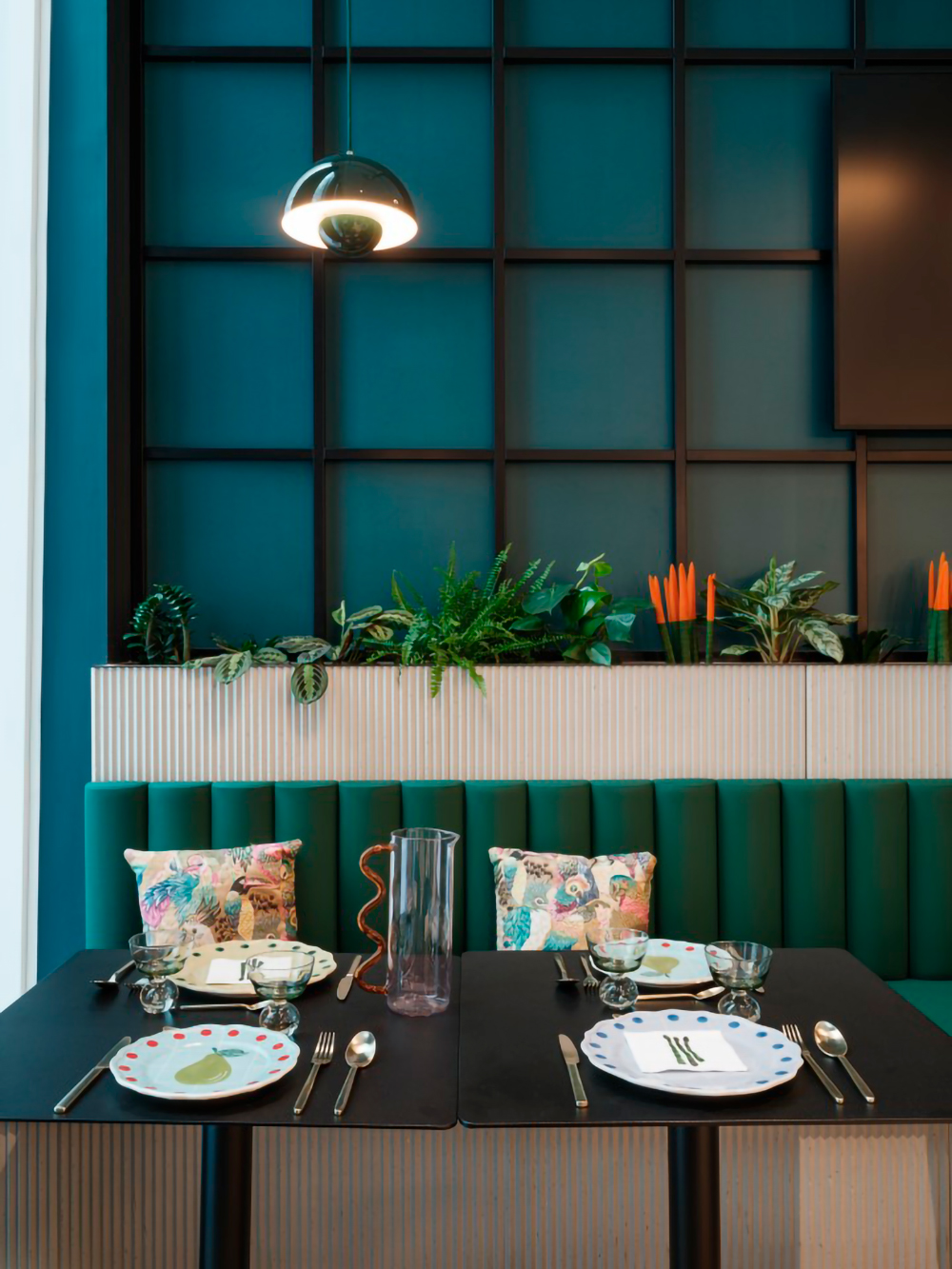 Une salle à manger moderne comprend une table dressée pour deux personnes avec des assiettes décoratives et des couverts. Derrière la table se trouve un banc vert rembourré orné de coussins fleuris. Une composition végétale et une suspension d'Ibis_KAI rehaussent l'ambiance sur un mur lambrissé bleu sarcelle foncé.