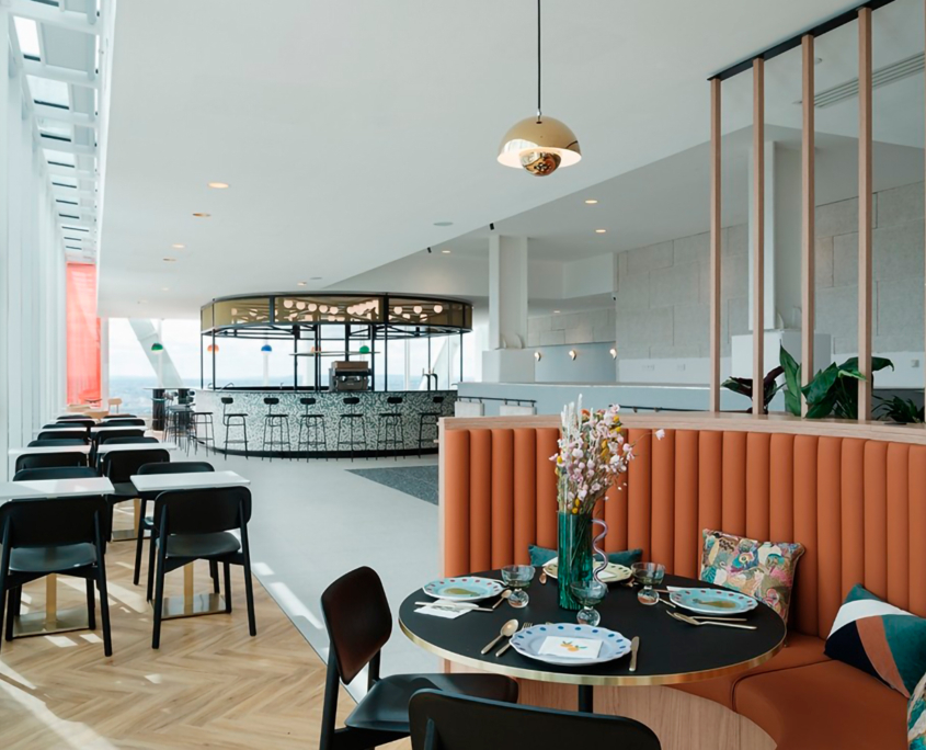 Un intérieur de restaurant moderne avec une table ronde dressée avec des assiettes et des verres, entourée de sièges rembourrés orange et de chaises noires. L'espace est lumineux, la lumière naturelle pénètre par de grandes fenêtres et l'élégant bar Ibis_KAI est visible en arrière-plan.