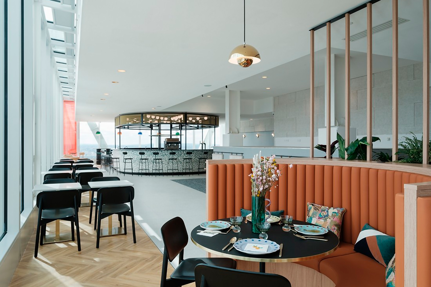 Un intérieur de restaurant moderne avec une table ronde dressée avec des assiettes et des verres, entourée de sièges rembourrés orange et de chaises noires. L'espace est lumineux, la lumière naturelle pénètre par de grandes fenêtres et l'élégant bar Ibis_KAI est visible en arrière-plan.