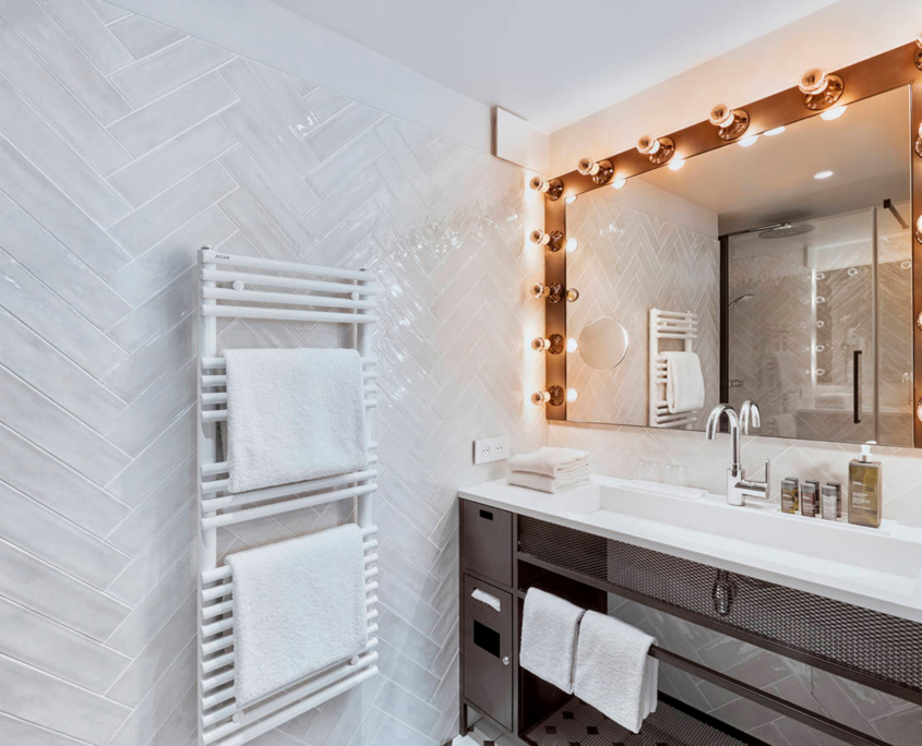 Une salle de bain moderne avec un mur carrelé en chevrons, un sèche-serviettes blanc avec des serviettes pliées et un grand miroir de courtoisie avec des rangées d'ampoules à gauche et à droite. Le meuble-lavabo, imprégné de l'élégance Ibis_KAI, est doté d'un comptoir blanc, d'un robinet chromé et de plusieurs bouteilles de toilette.