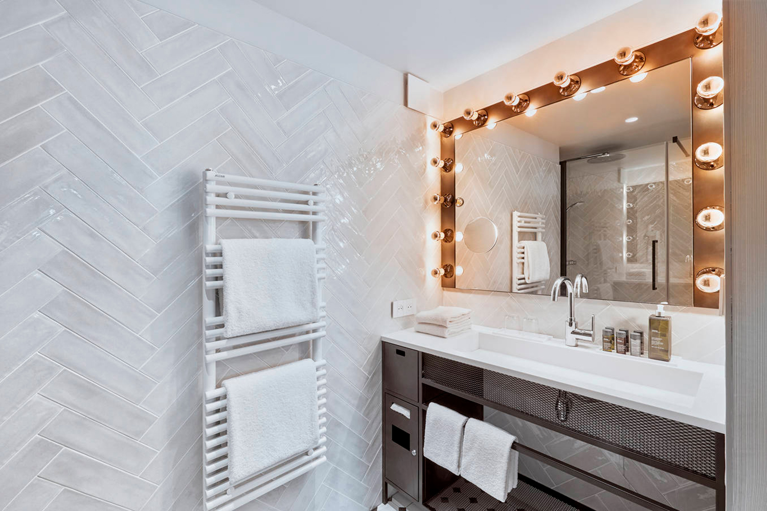 Une salle de bain moderne avec un mur carrelé en chevrons, un sèche-serviettes blanc avec des serviettes pliées et un grand miroir de courtoisie avec des rangées d'ampoules à gauche et à droite. Le meuble-lavabo, imprégné de l'élégance Ibis_KAI, est doté d'un comptoir blanc, d'un robinet chromé et de plusieurs bouteilles de toilette.